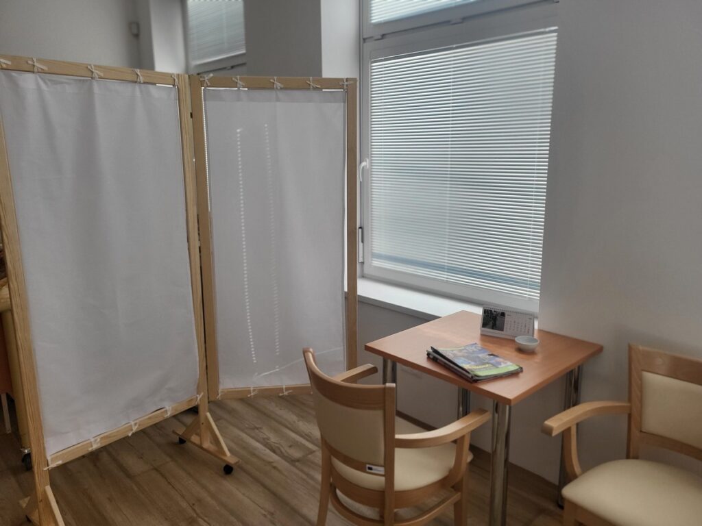 Malá miestnosť s dreveným stolom, dvoma stoličkami, zástenou a oknom so zatiahnutými žalúziami. Na stole sú časopisy a tanier. Podlaha je drevená a atmosféra je tichá a klinická.