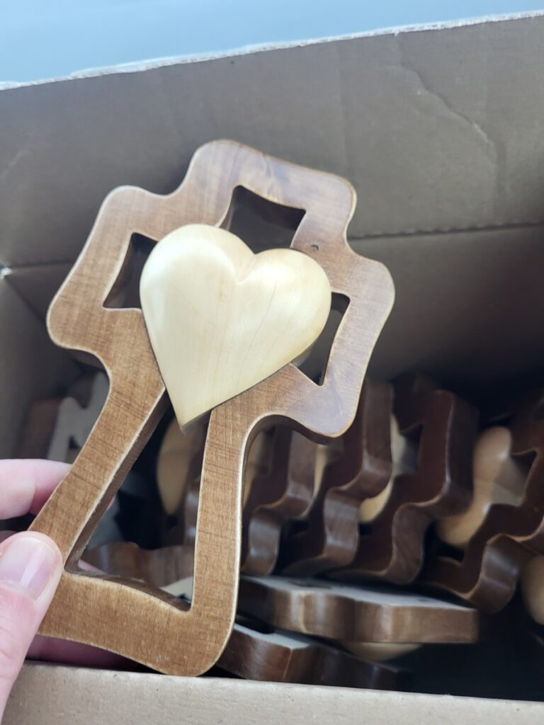 Ruka drží drevený predmet v tvare kríža s vyvýšeným srdcom v strede nad škatuľou naplnenou podobnými drevenými predmetmi v tvare kríža a srdca.