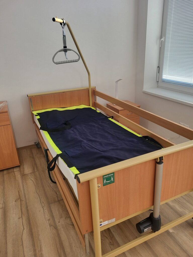 Drevená nemocničná posteľ so zábradlím, hrazda nad hlavou a tmavomodré prestieradlo na zdvíhanie pacientov so žltými okrajmi; izba má svetlé steny, okno so žalúziami a drevenú podlahu.