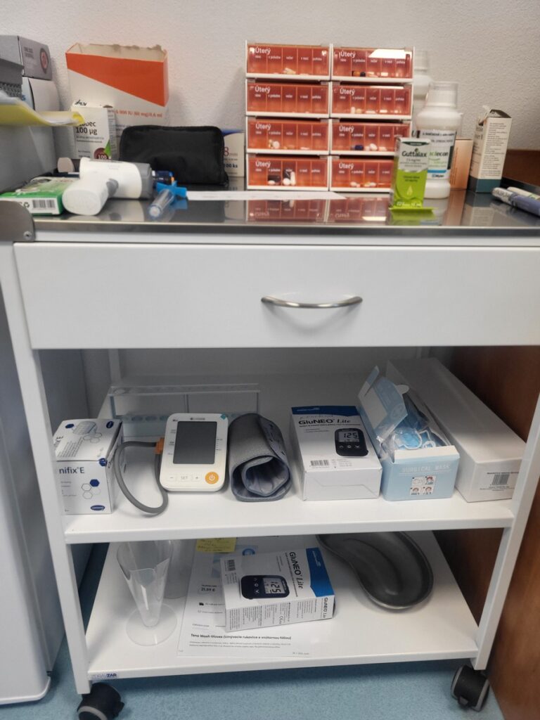 Lekársky vozík s rôznymi potrebami: lieky a nádoby na vrchu, tlakomer, senzor GlucoMen, škatule a hadičky na strednej polici a ďalšie lekárske vybavenie a škatule na spodnej polici.