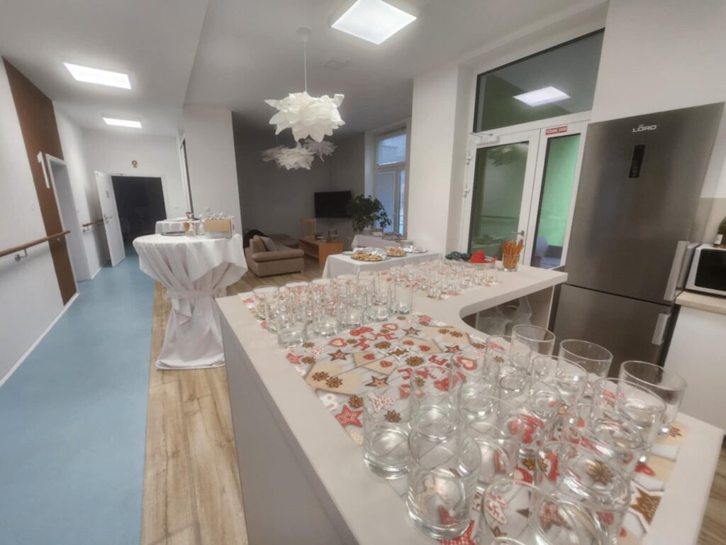 Miestnosť pripravená na podujatie s radmi prázdnych pohárov na slávnostnom obruse, stolom s občerstvením, koktailovými stolíkmi, pohovkou a chladničkou v pozadí. Priestor je svetlý s modernou výzdobou.