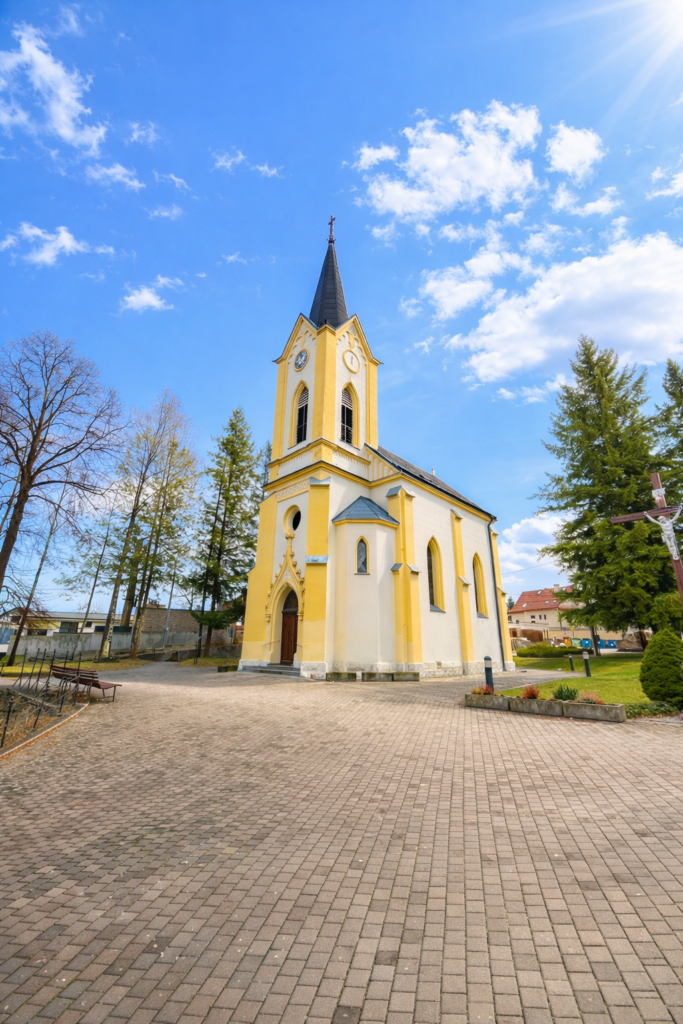 Žltý kostol s vysokou špicatou vežou a hodinami stojí na vydláždenom námestí obklopenom stromami pod jasne modrou oblohou s roztrúsenými mrakmi.