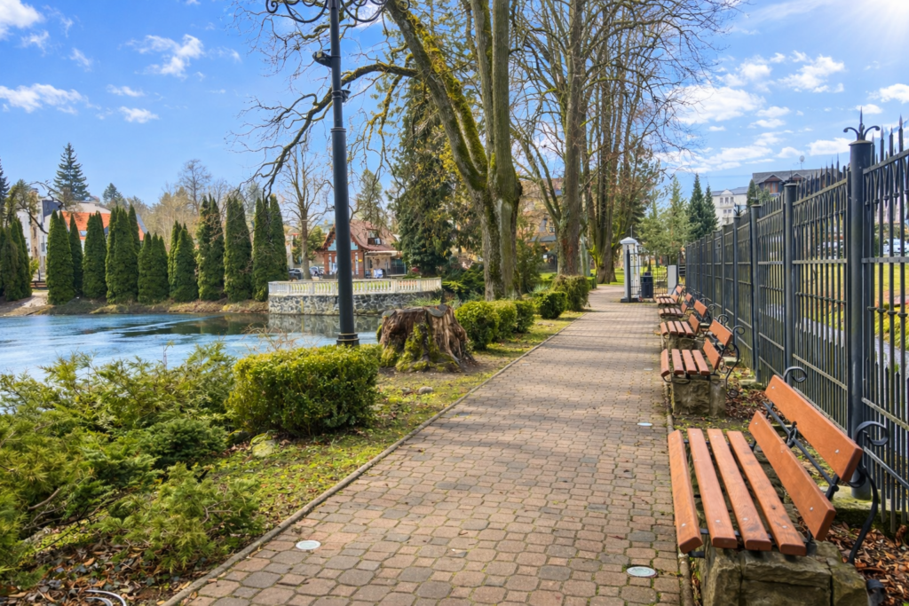 Vydláždený parkový chodník s drevenými lavičkami vedie popri rieke, ktorú lemujú stromy a kovové oplotenie. Cestu lemujú kríky a v pozadí sú pod modrou oblohou viditeľné domy a vždyzelené stromy.